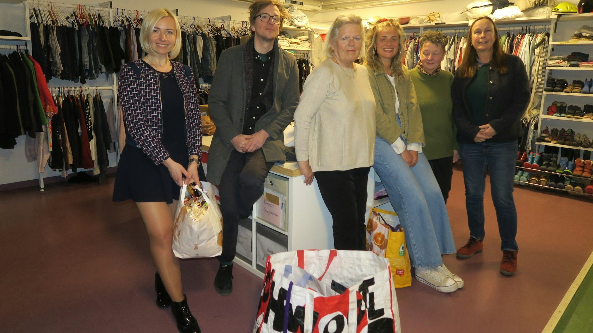 Sechs Menschen stehen nebeneinander an einem Tisch. Hinter ihnen hängen Kleider an den Wänden eines Kellerraums.