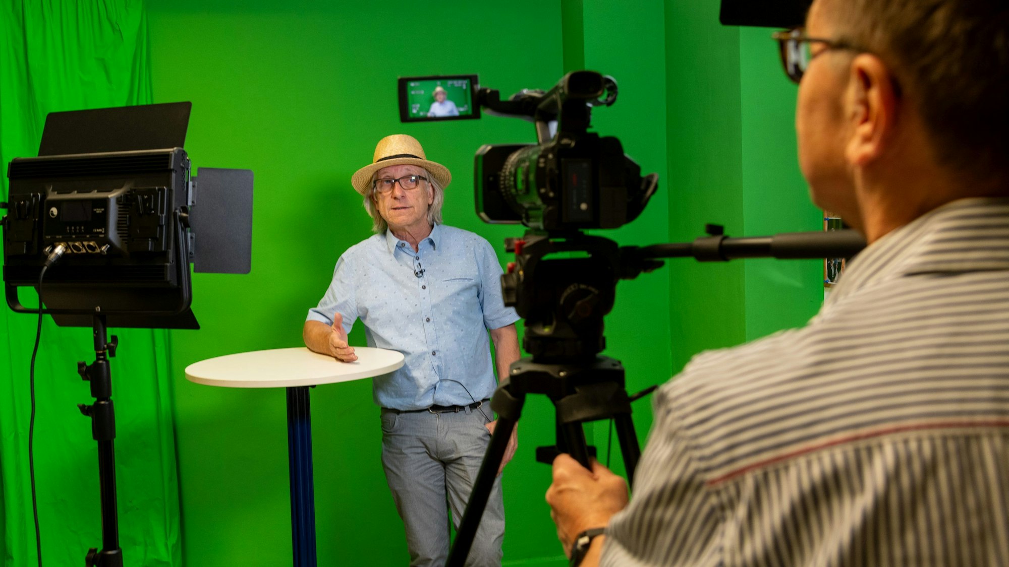 Ein Mann steht an einem Stehtisch vor einer grünen Wand. Ein anderer Mann filmt ihn.