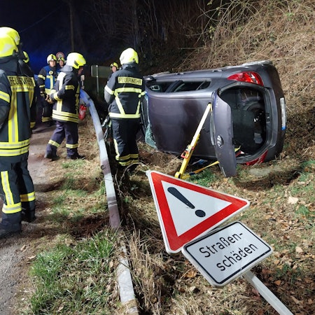 Ein Auto liegt am Straßenrand auf der Seite. Links Feuerwehrleute. Im Vordergrund ein Straßenschild mit der Aufschrift „Straßenschäden“.