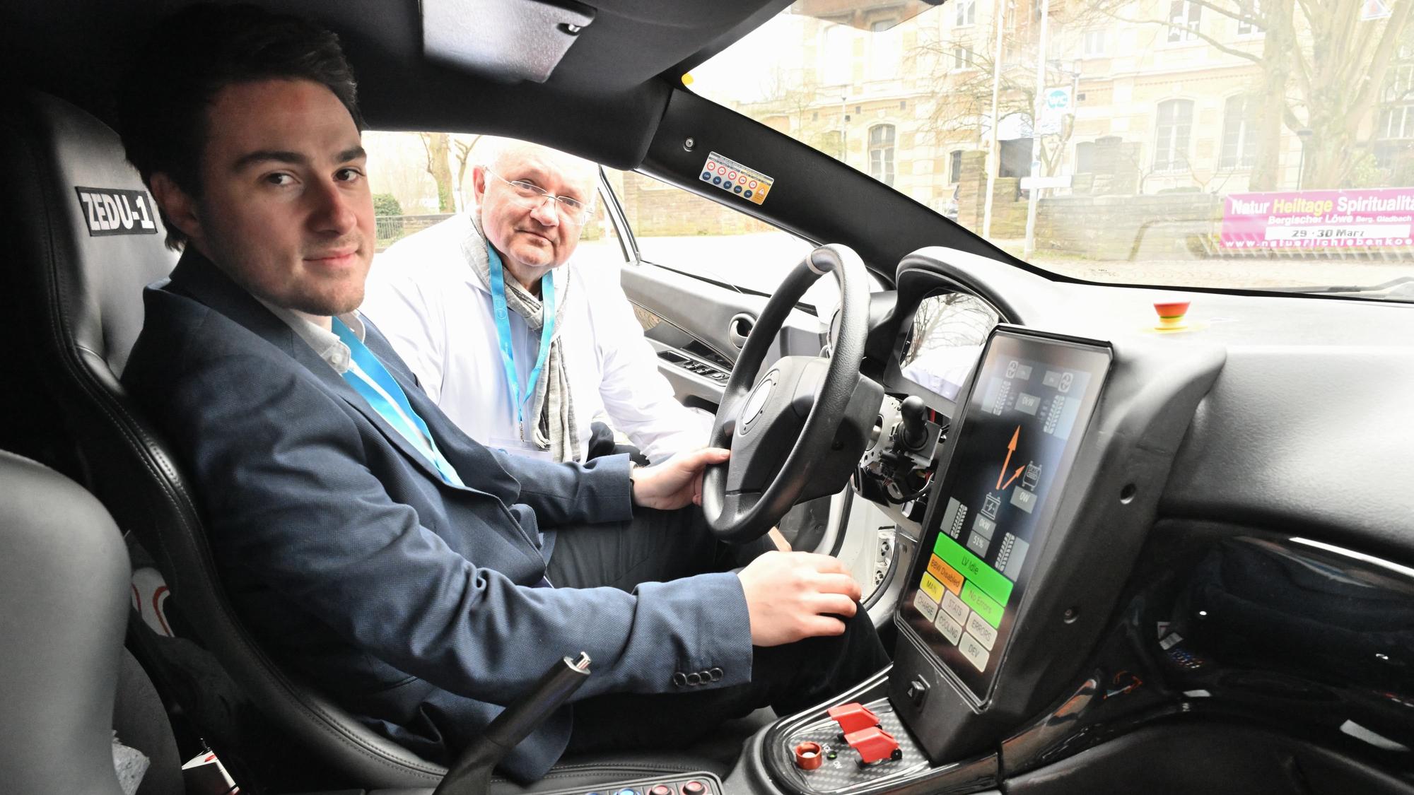 In dem Auto mit vielen Knöpfen und großem Display sitzt ein Mann, ein anderer schaut zur offenen Fahrertür herein.