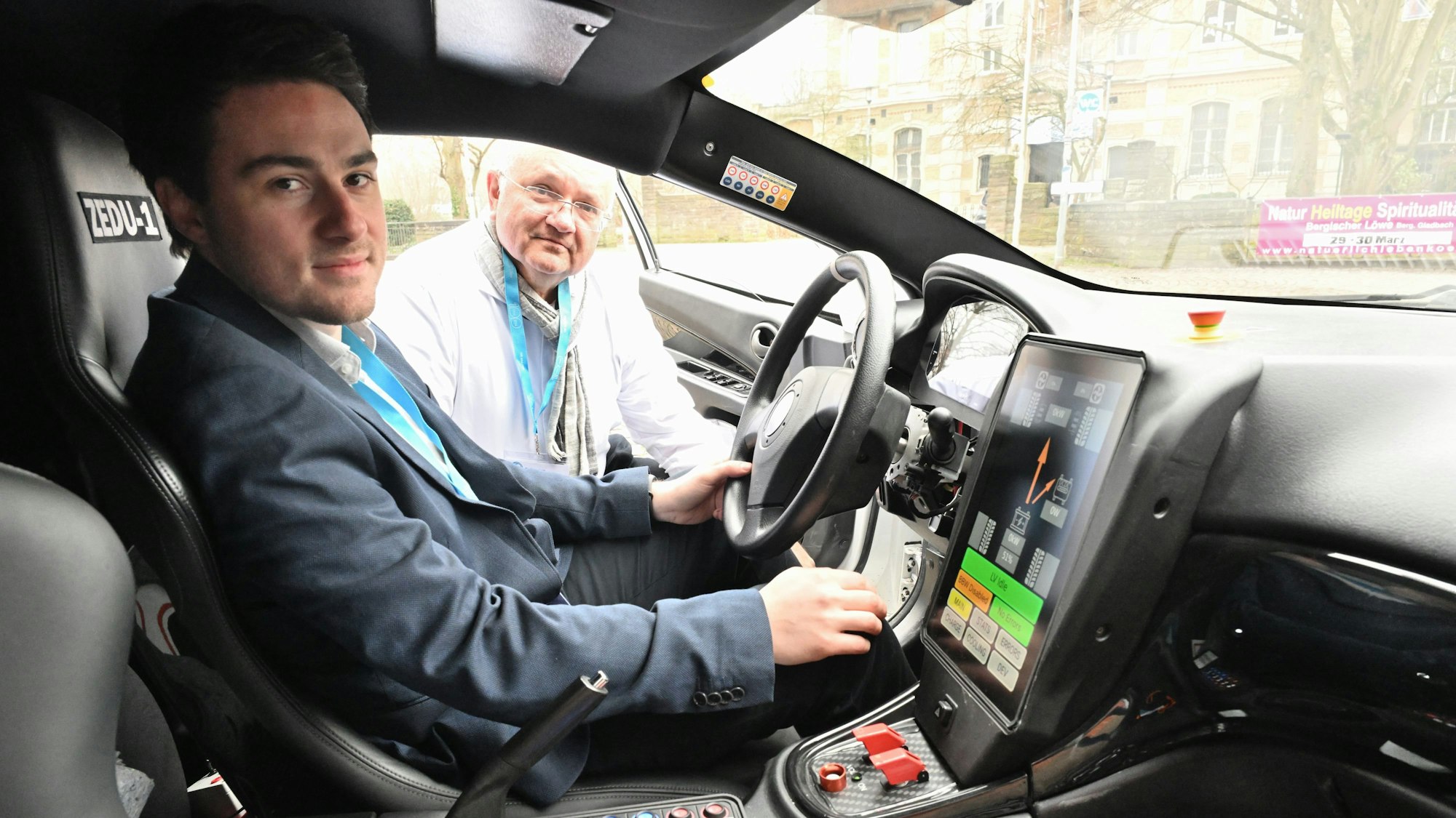 In dem Auto mit vielen Knöpfen und großem Display sitzt ein Mann, ein anderer schaut zur offenen Fahrertür herein.