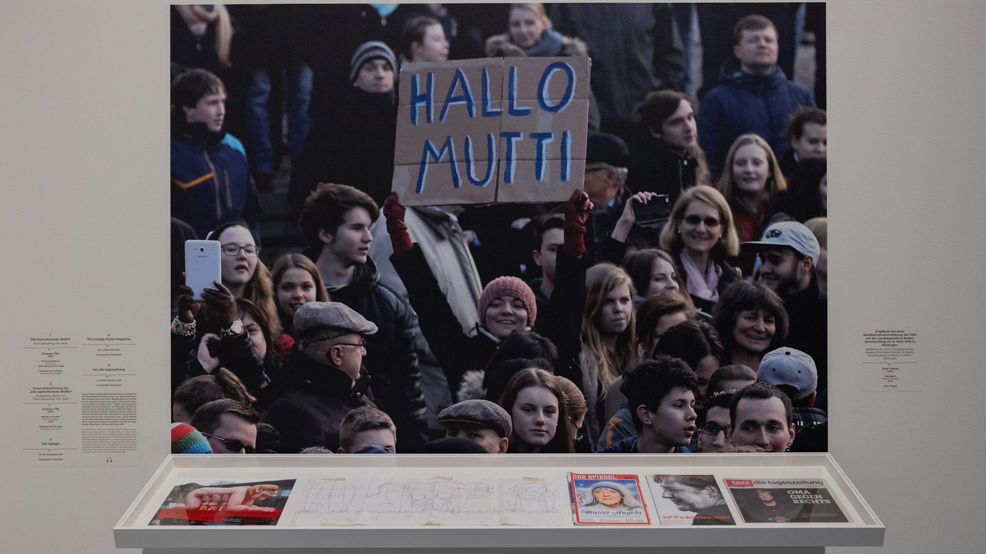 PRODUKTION - 10.03.2025, Nordrhein-Westfalen, Düsseldorf: Zeitschriftentitel, eine Skizze eines Karnevalswagens, die die damalige Bundeskanzlerin Merkel zeigen und ein Pressefoto einer CDU-Wahlkampfveranstaltung von Sean Gallup, auf dem ein Schild mit der Aufschrift ·Hallo Mutti· gezeigt wird, sind in der Ausstellung ·Mama. Von Maria bis Merkel· im Museum Kunstpalast zu sehen. Foto: Rolf Vennenbernd/dpa - ACHTUNG: Nur zur redaktionellen Verwendung im Zusammenhang mit der aktuellen Berichterstattung über die genannte Ausstellung und nur mit vollständiger Nennung des vorstehenden Credits. +++ dpa-Bildfunk +++