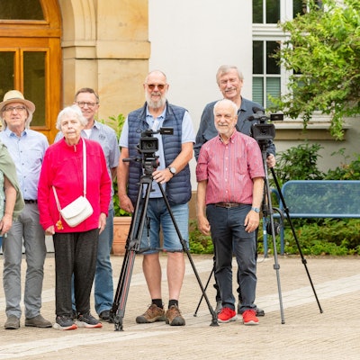 Eine Gruppe älterer Menschen steht vor einem Gebäude. Sie haben zwei Kameras auf Stativen dabei.