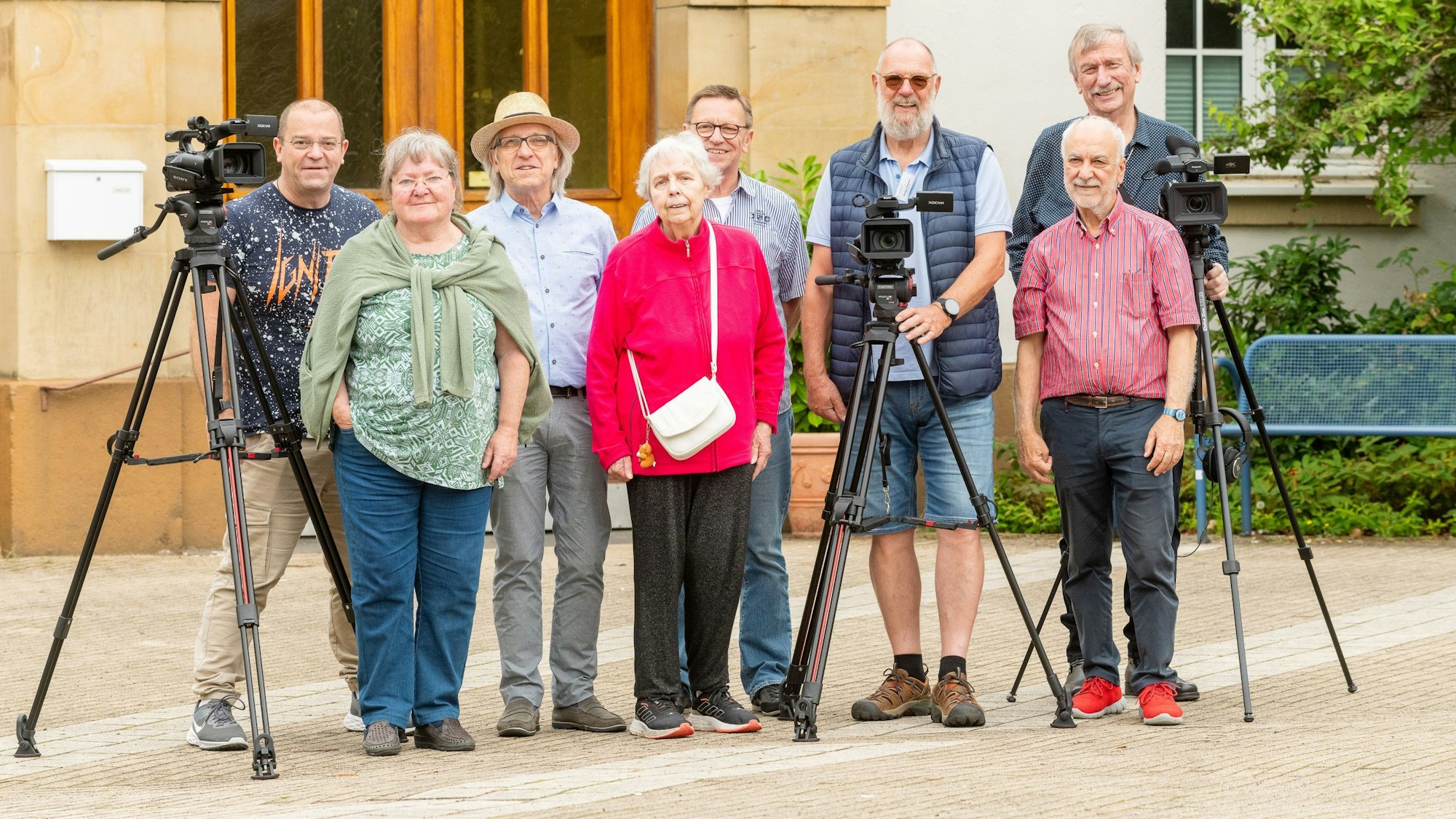 Eine Gruppe älterer Menschen steht vor einem Gebäude. Sie haben zwei Kameras auf Stativen dabei.