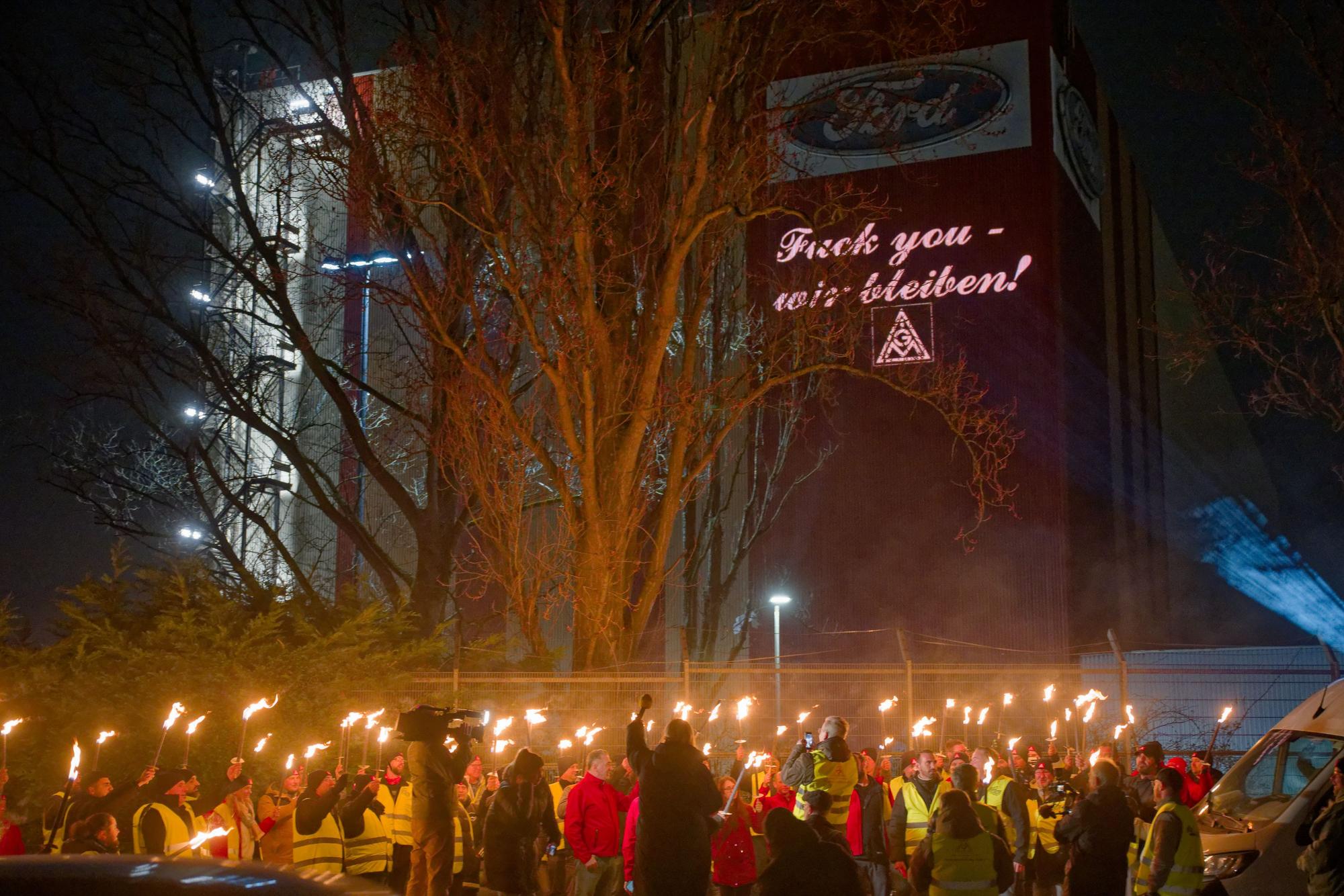 Ford-Mitarbeitende stehen mit Fackeln auf dem Gehweg vor einem Gebäude der Fordwerke, auf dem die Worte "Fuck you - wir bleiben" projiziert wird.