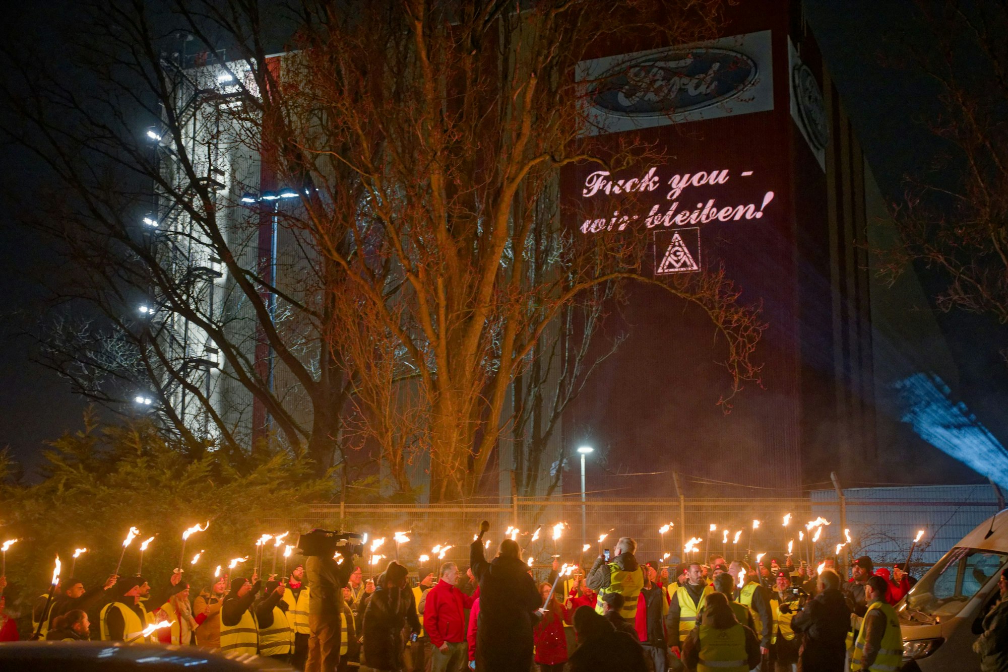 Ford-Mitarbeitende stehen mit Fackeln auf dem Gehweg vor einem Gebäude der Fordwerke, auf dem die Worte "Fuck you - wir bleiben" projiziert wird.