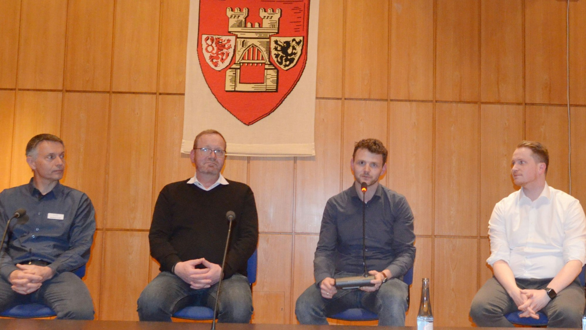 Vier Männer sitzen nebeneinander auf Stühlen, über ihnen an der Wand das Wappen der Stadt Euskirchen.
