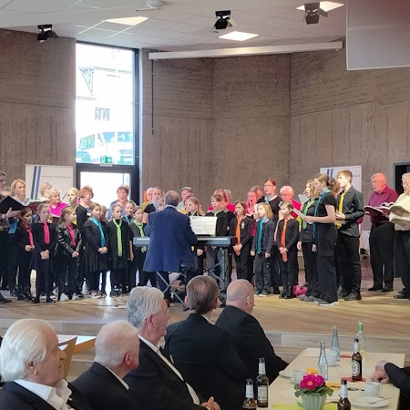 Kinder, Frauen und Männer singen gemeinsam auf einer Bühne.