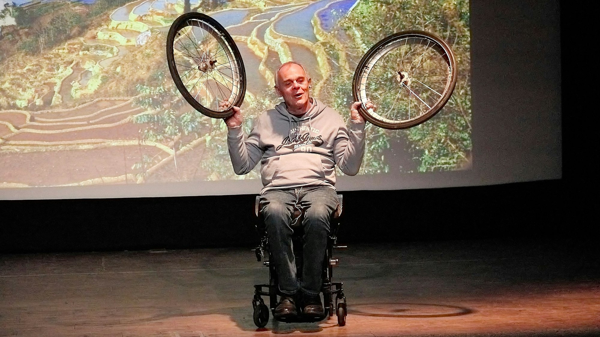 Andreas Pröve sitzt auf einer Bühne in seinem Rollstuhl und hält beide Räder in die Höhe. Im Hintergrund ist eine Leinwand zu erkennen.