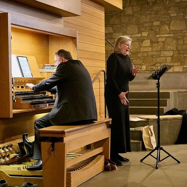 Ein Mann in schwarzem Anzug sitzt an einer Orgel. Neben ihm steht eine Frau, ebenfalls schwarz gekleidet mit blonden Haaren, und singt.
