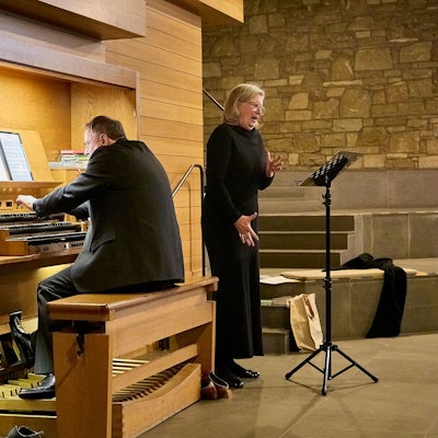 Ein Mann in schwarzem Anzug sitzt an einer Orgel. Neben ihm steht eine Frau, ebenfalls schwarz gekleidet mit blonden Haaren, und singt.