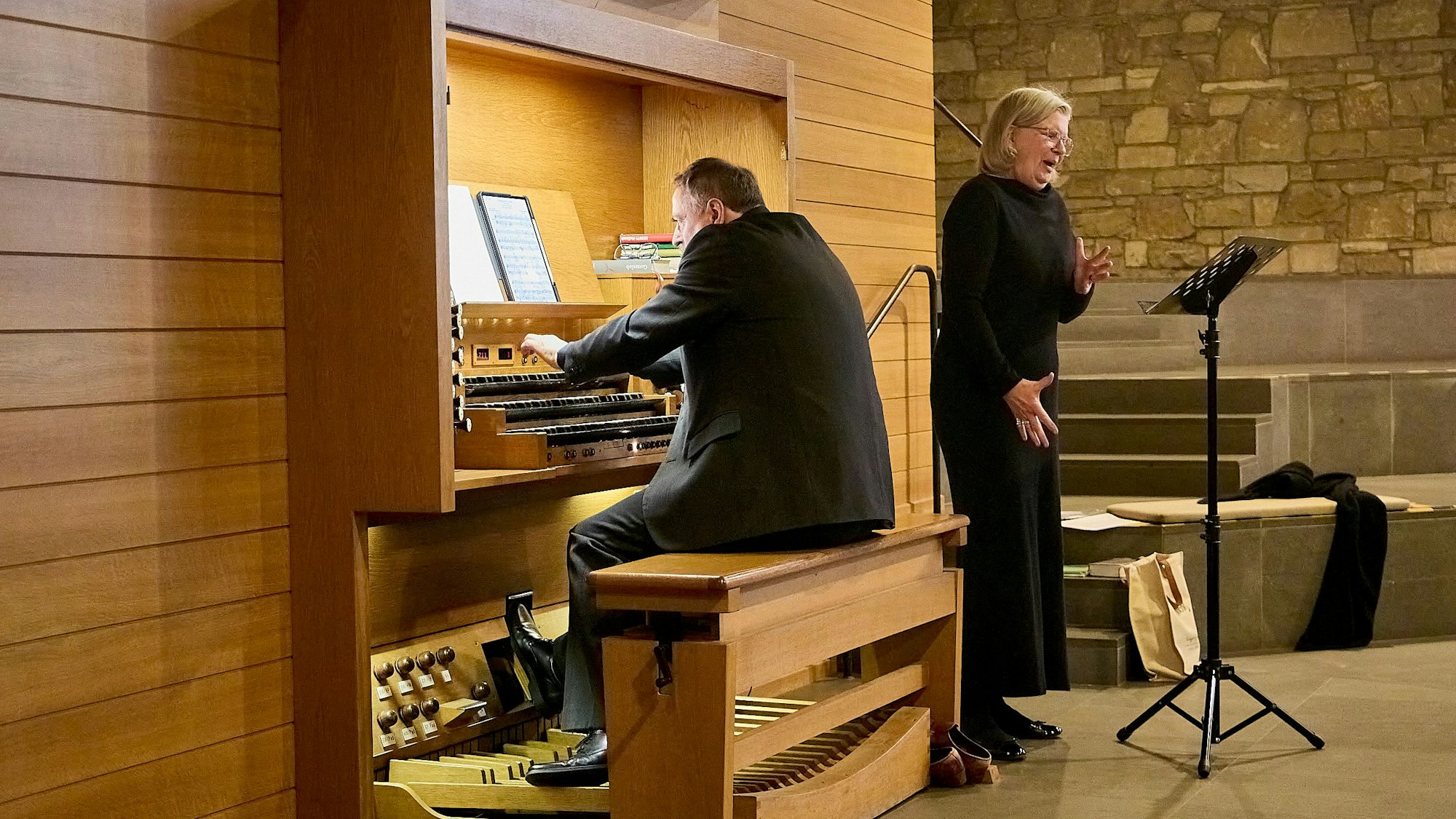 Ein Mann in schwarzem Anzug sitzt an einer Orgel. Neben ihm steht eine Frau, ebenfalls schwarz gekleidet mit blonden Haaren, und singt.