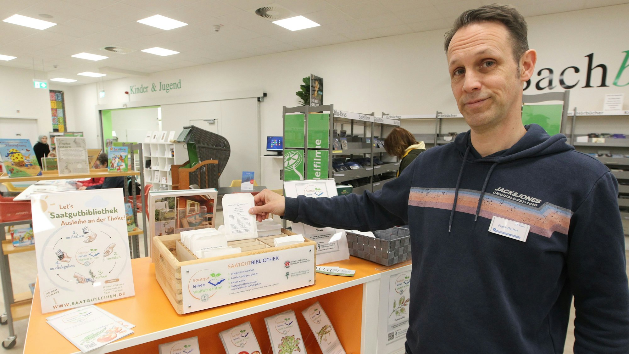 Ein Mann steht an einem Regal in einer Bibliothek. Aus einem hölzernen Kasten hält er eine kleine Papiertüte hoch.