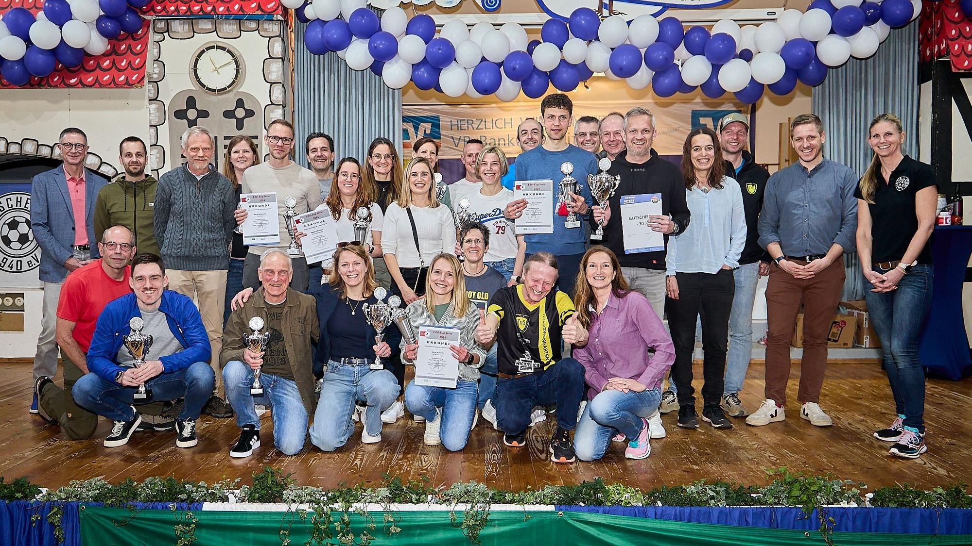 Zahlreiche Frauen und Männer stehen und hocken auf der Bühne im Dorfsaal in Scheven. Sie halten Pokale und Urkunden in den Händen, die sie beim Eifelcup gewonnen haben.