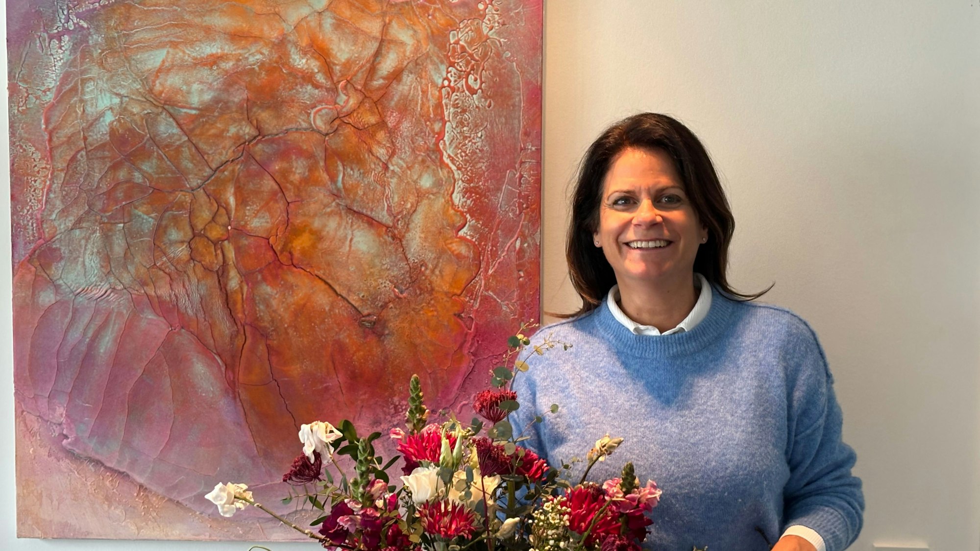 Unten mittig im Bild steht ein Blumenstrauß. Dahinter steht eine Frau neben einem Bild, das an der Wand hängt, und lächelt in die Kamera.