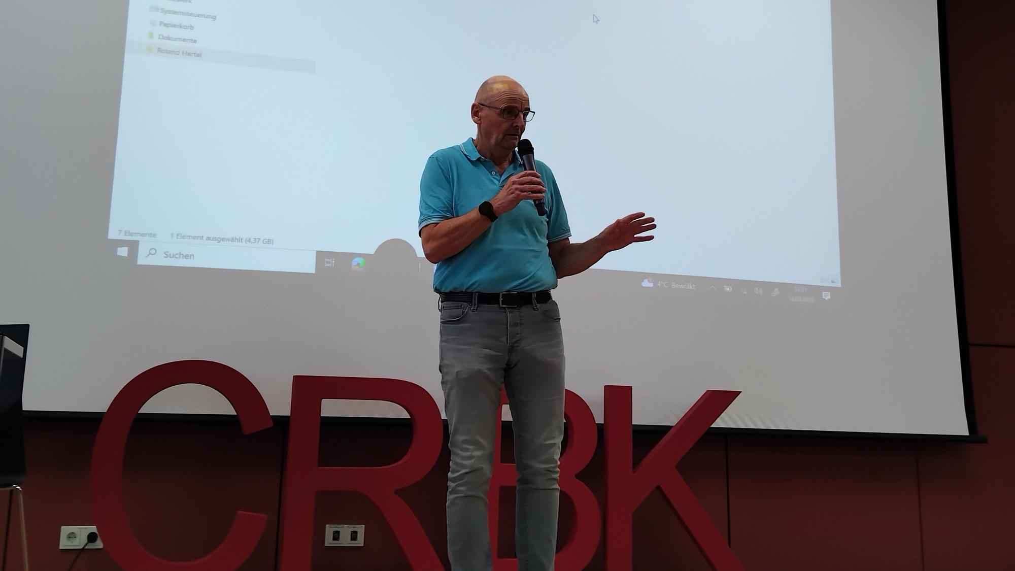 Ein Mann steht auf einer Bühne und spricht gestikulierend in ein Mikrophon.