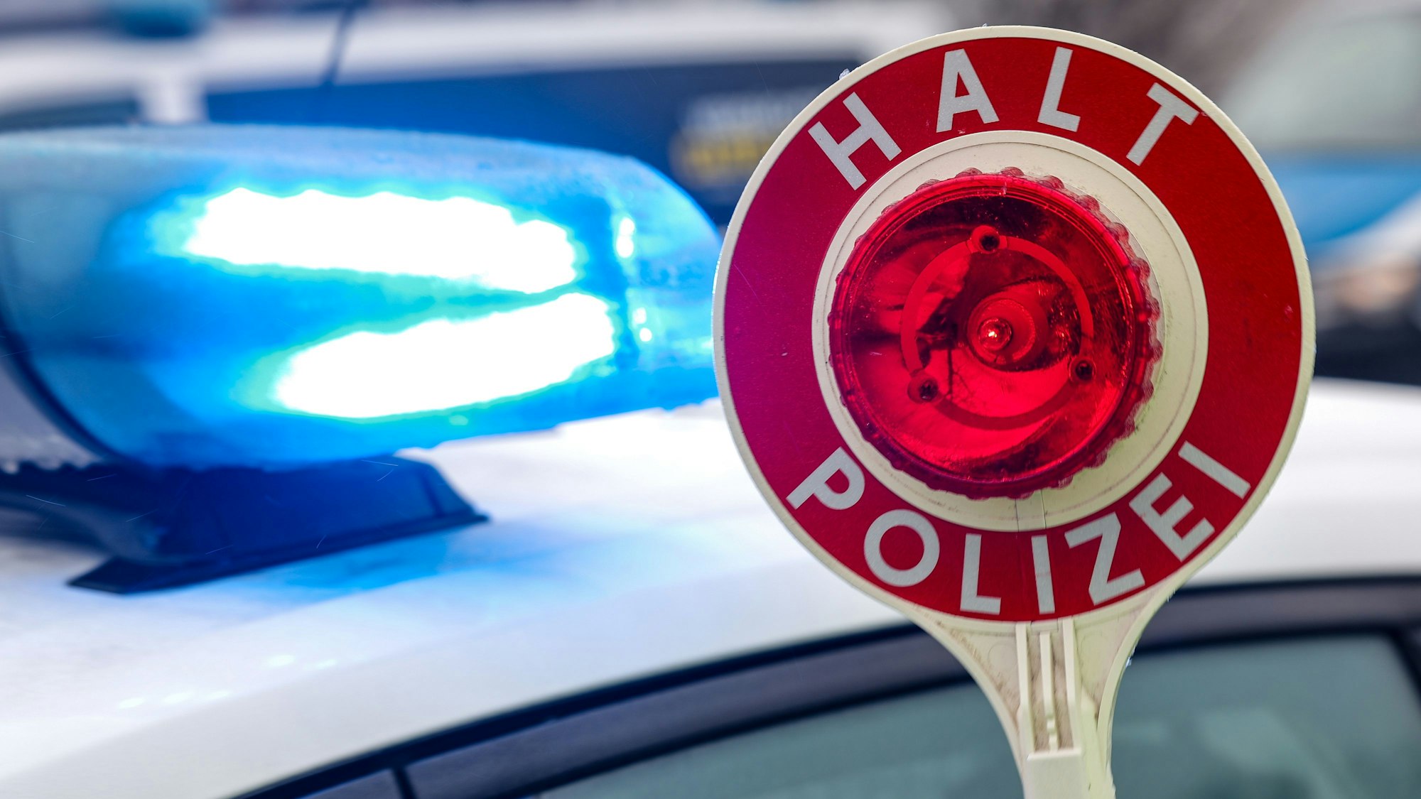 Eine Kelle mit der Aufschrift „Halt Polizei“ ist vor einem Streifenwagen zu sehen, dessen Blaulicht eingeschaltet ist.