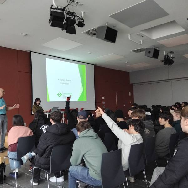 Eine Gruppe Jugendlicher und junger Erwachsener sitzt in einer Aula, zwei Leute haben die Hände gehoben. Ein älterer Mann steht gestikulierend vor ihnen. Auf einer Leinwand vorne steht „Häusliche Gewalt - Probleme?“