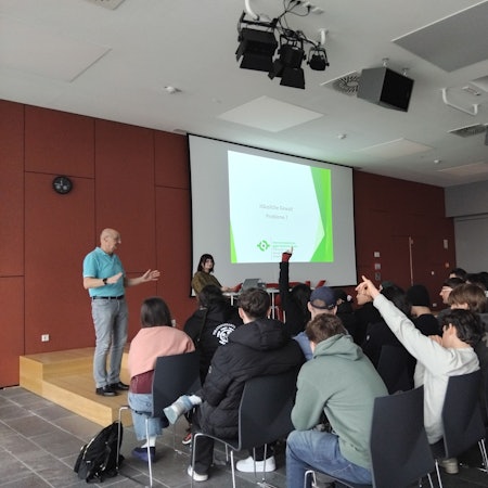 Eine Gruppe Jugendlicher und junger Erwachsener sitzt in einer Aula, zwei Leute haben die Hände gehoben. Ein älterer Mann steht gestikulierend vor ihnen. Auf einer Leinwand vorne steht „Häusliche Gewalt - Probleme?“