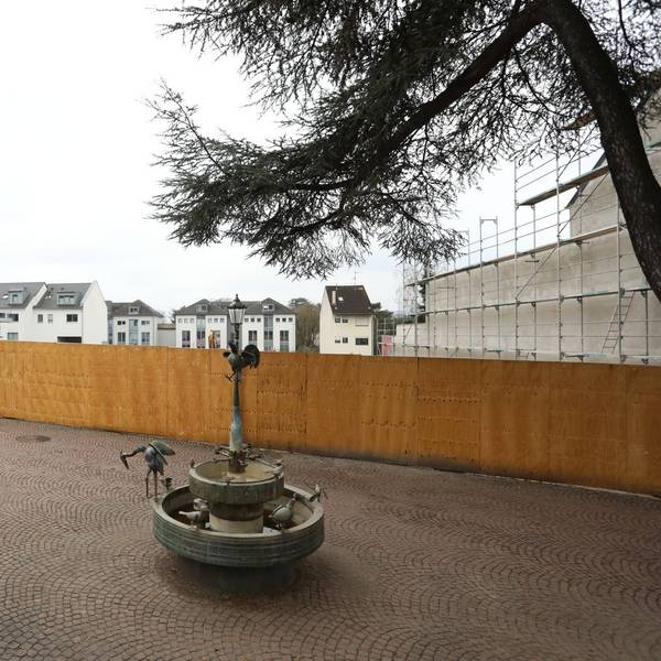 Ein Brunnen und einige Tische und Stühle stehen in der Fußgängerzone, im Hintergrund steht ein großer Bauzaun.