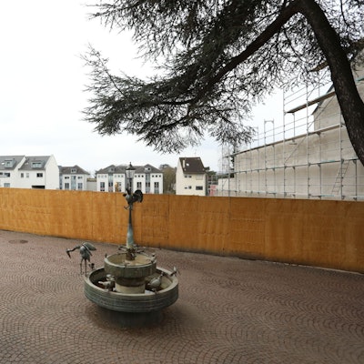 Ein Brunnen und einige Tische und Stühle stehen in der Fußgängerzone, im Hintergrund steht ein großer Bauzaun.