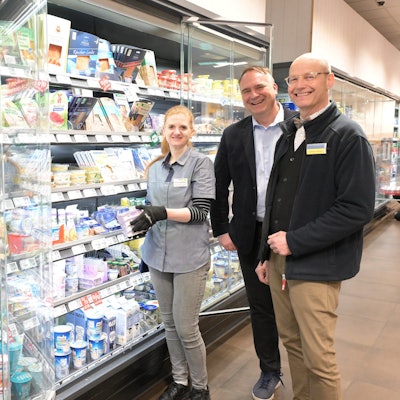 Eine Frau und zwei Männer stehen in einem Supermarkt vor einem Kühlregal mit Milchprodukten.