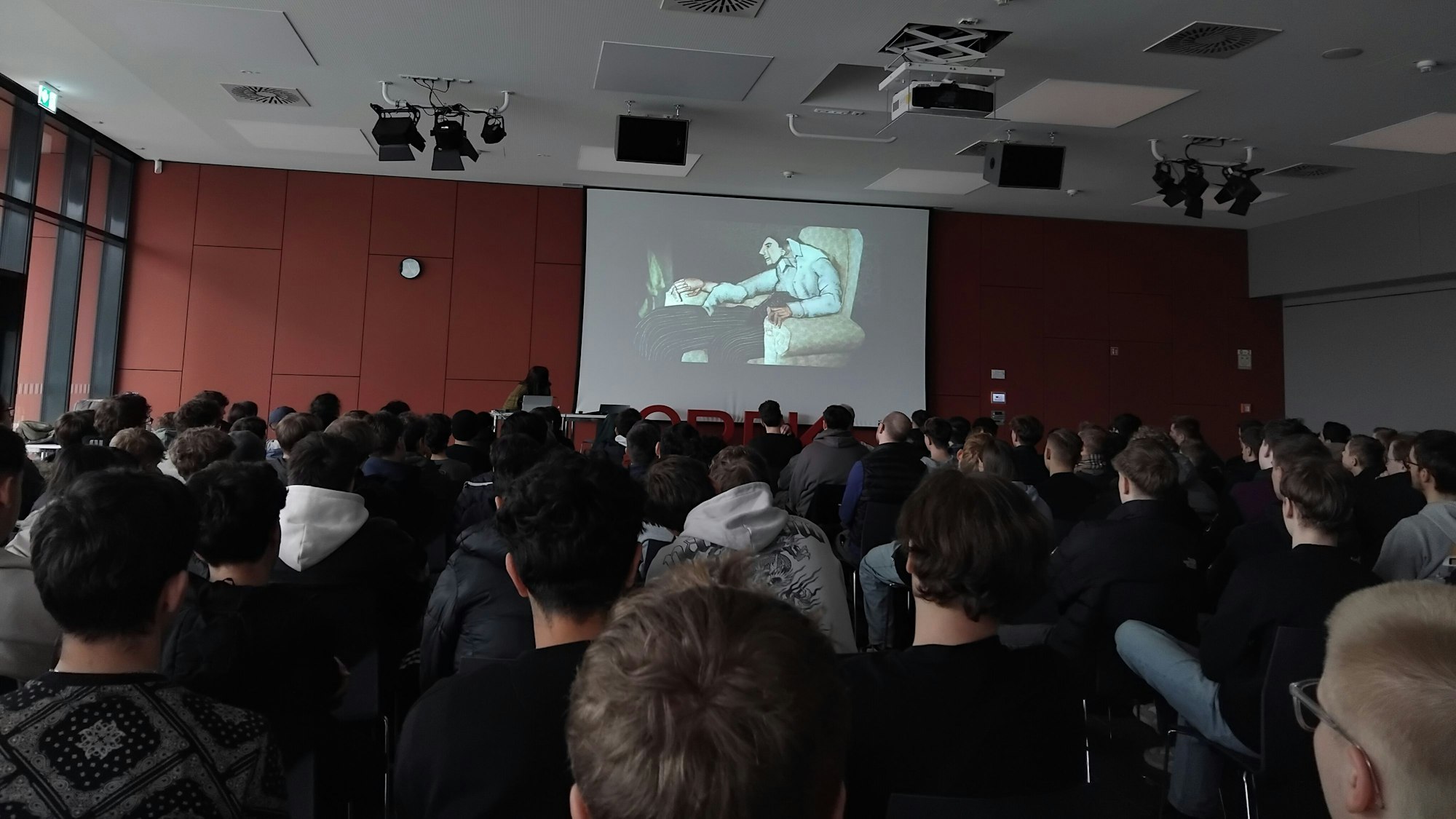 Zahlreiche junge Leute, vorwiegend männlich, sitzen in einer Aula, vorne wird auf einer Leinwand ein Film gezeigt.