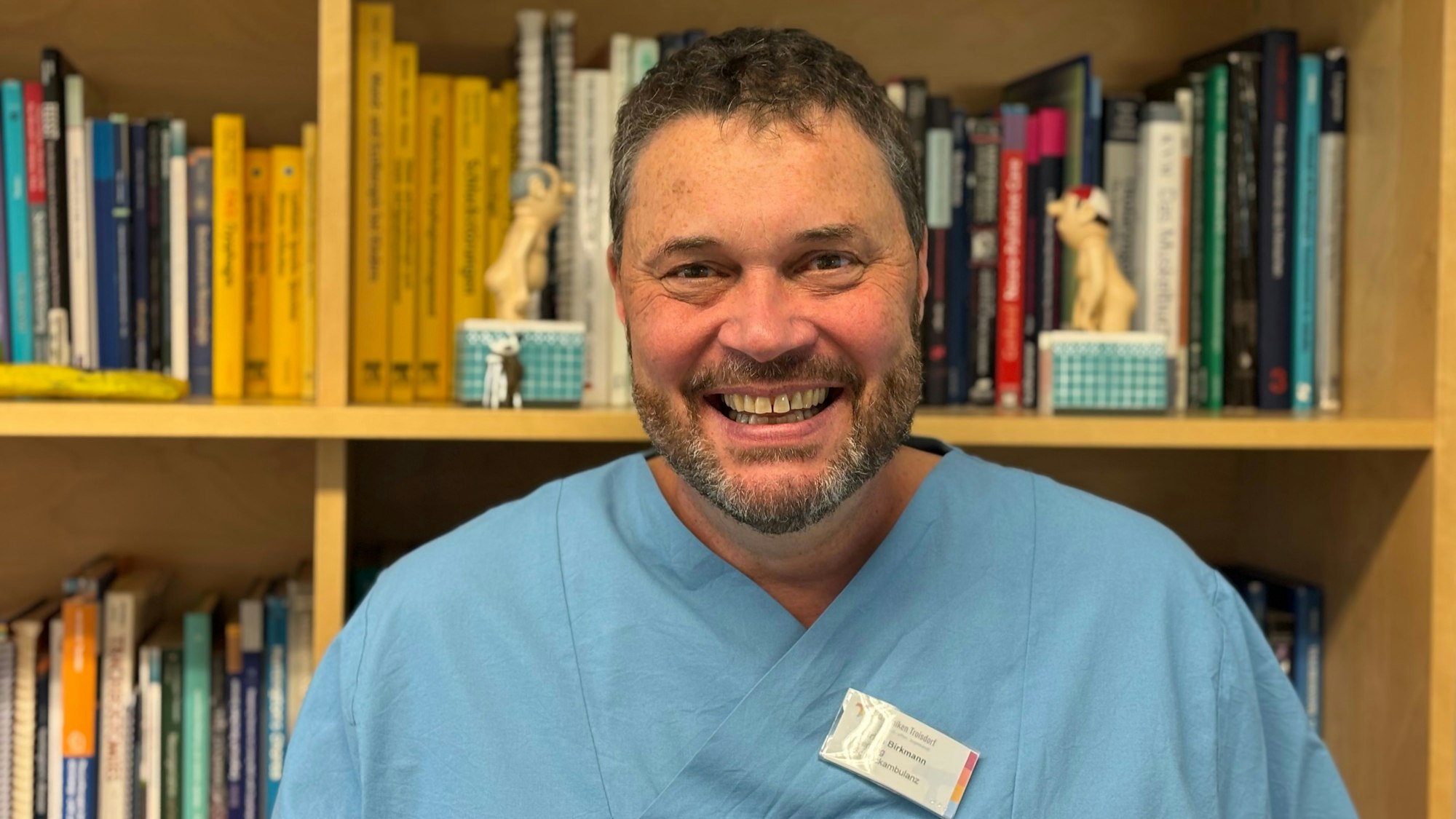 Ein Mann im blauen Ärztekittel lächelt breit in die Kamera. Im Hintergrund befindet sich ein unscharf gestelltes Buchregal mit zwei Loriotfiguren.