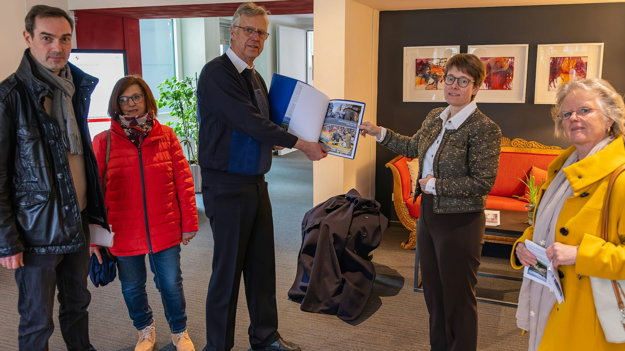 Eine Gruppe Menschen stehen in einem Raum. Ein Mann überreicht einer Frau Unterlagen.