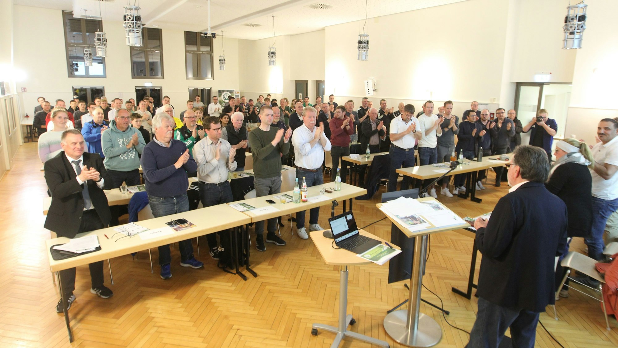 Teilnehmerinnen und Teilnehmer einer Versammlung stehen an ihren Tischen und klatschen.