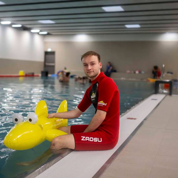 Ein Schwimmlehrer in rotem Neoprenanzug sitzt am Rand des Schwimmbackens der Schwimmschule Sharky in Weilerswist. Die Beine baumeln im Wasser. Er hält einen gelben Plastik-Hai fest.