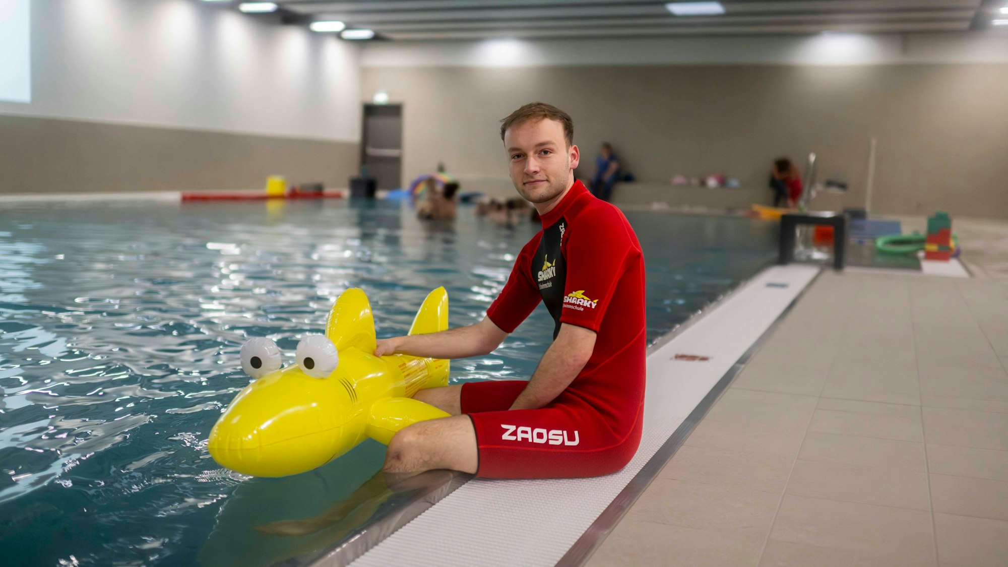 Ein Schwimmlehrer in rotem Neoprenanzug sitzt am Rand des Schwimmbackens der Schwimmschule Sharky in Weilerswist. Die Beine baumeln im Wasser. Er hält einen gelben Plastik-Hai fest.