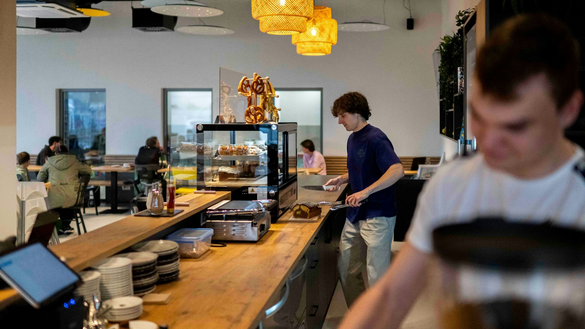 Zu der neuen Schwimmschule gehört auch ein Café. Zwei Männer arbeiten am Tresen, im Hintergrund sitzen Gäste an den Tischen.