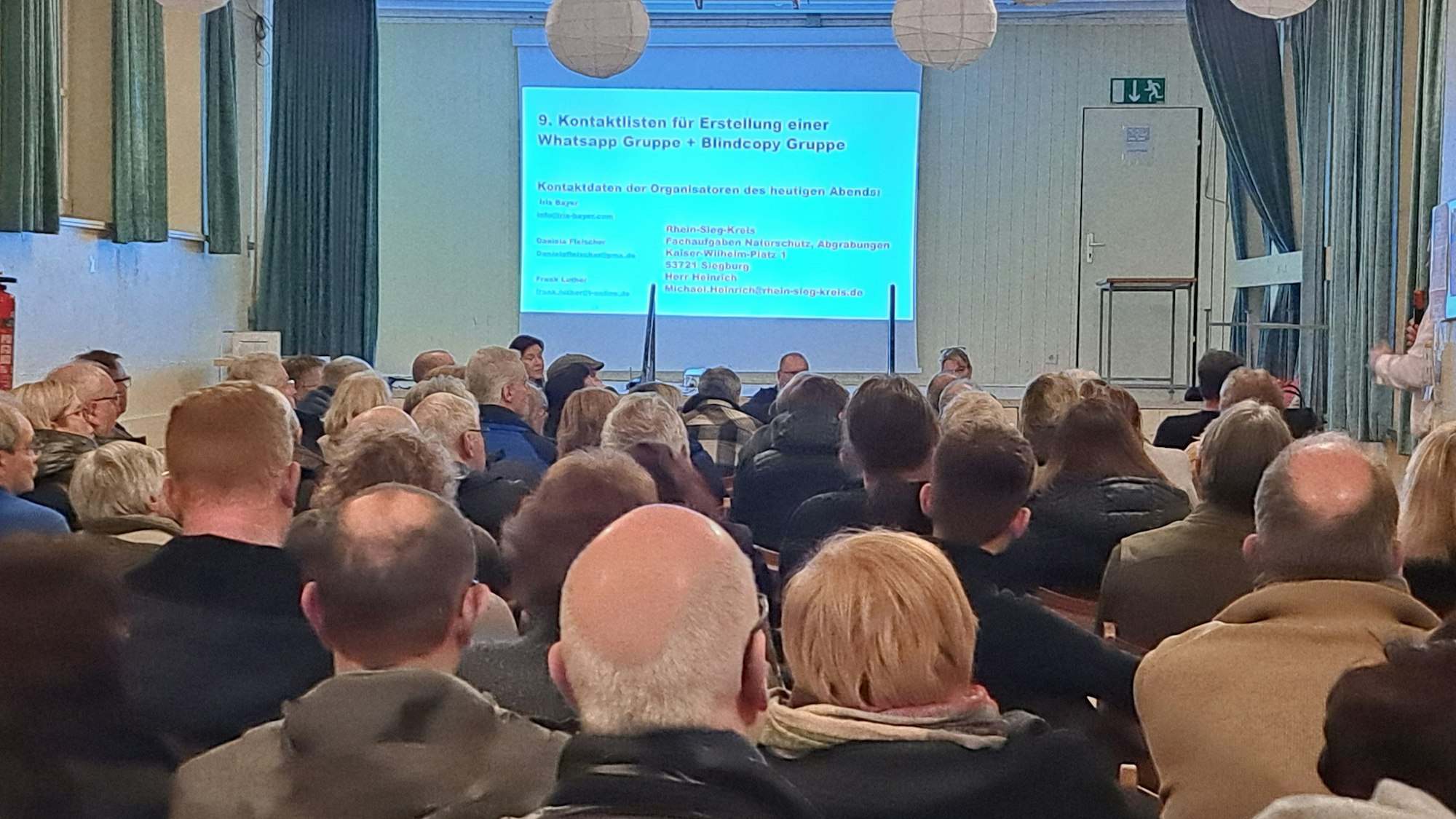 Männer und Frauen in einem Veranstaltungssaal schauen auf eine Projektion an der Wand des Raums.