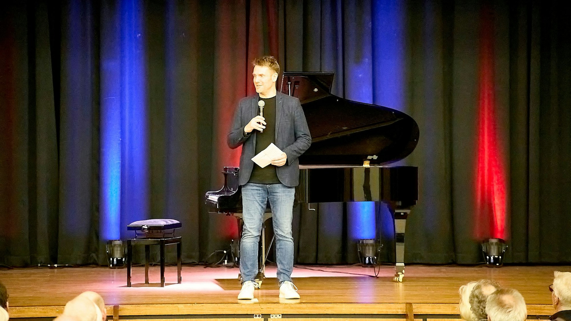 Alexander Joist steht auf der Bühne und spricht ins Mikrofon. Hinter ihm steht der Flügel.