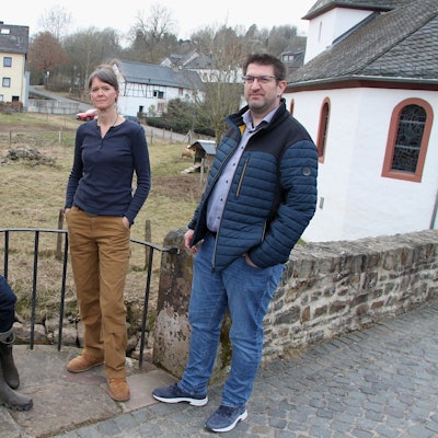 Zwei Männer und eine Frau stehen an einer alten Bruchsteinmauer, im Hintergrund sind Wiesen und Häuser zu sehen.