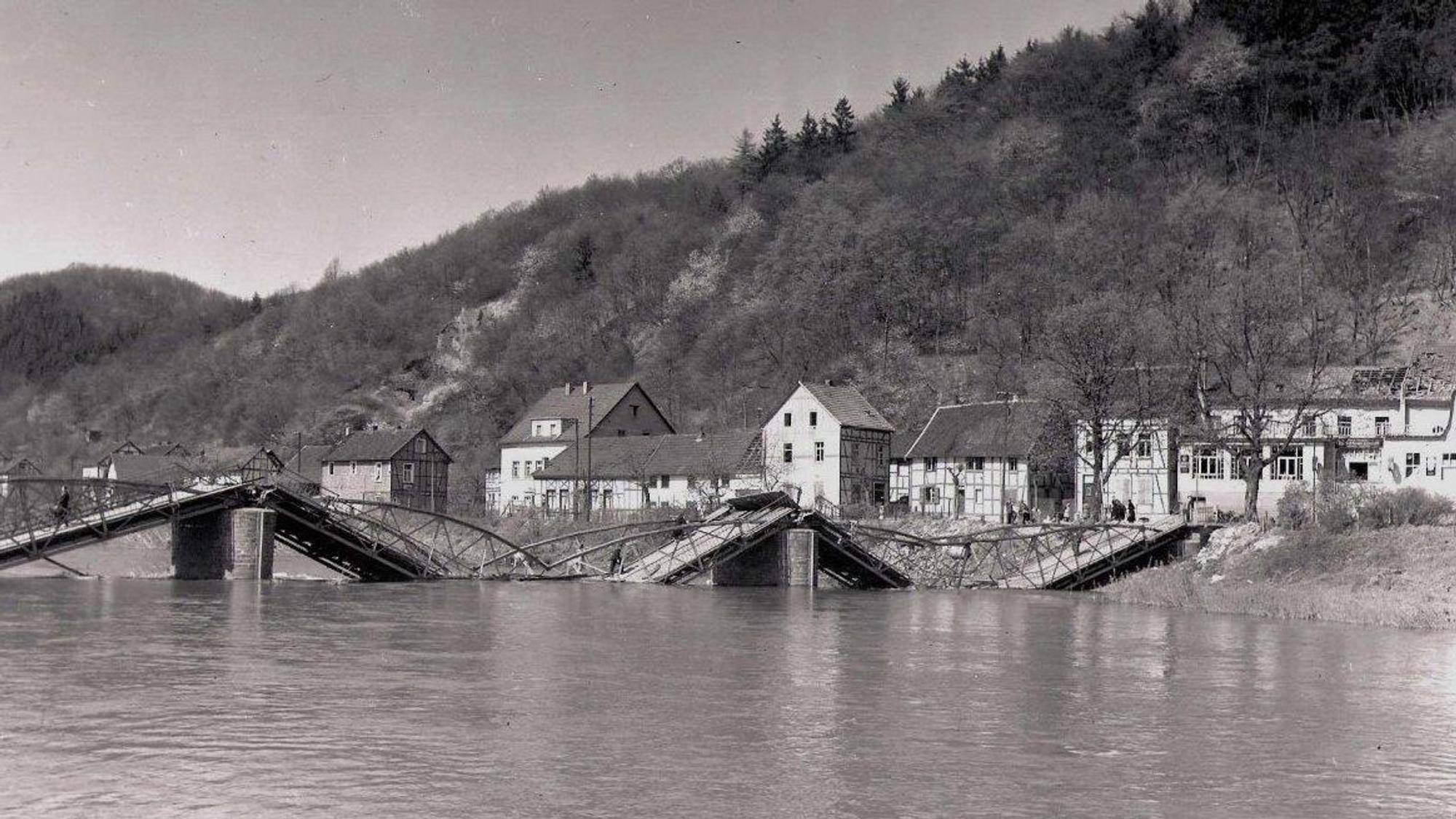 Schwarz-Weiß-Foto einer zerstörten Brücke; die Fahrbahnteile liegen im Fluss.