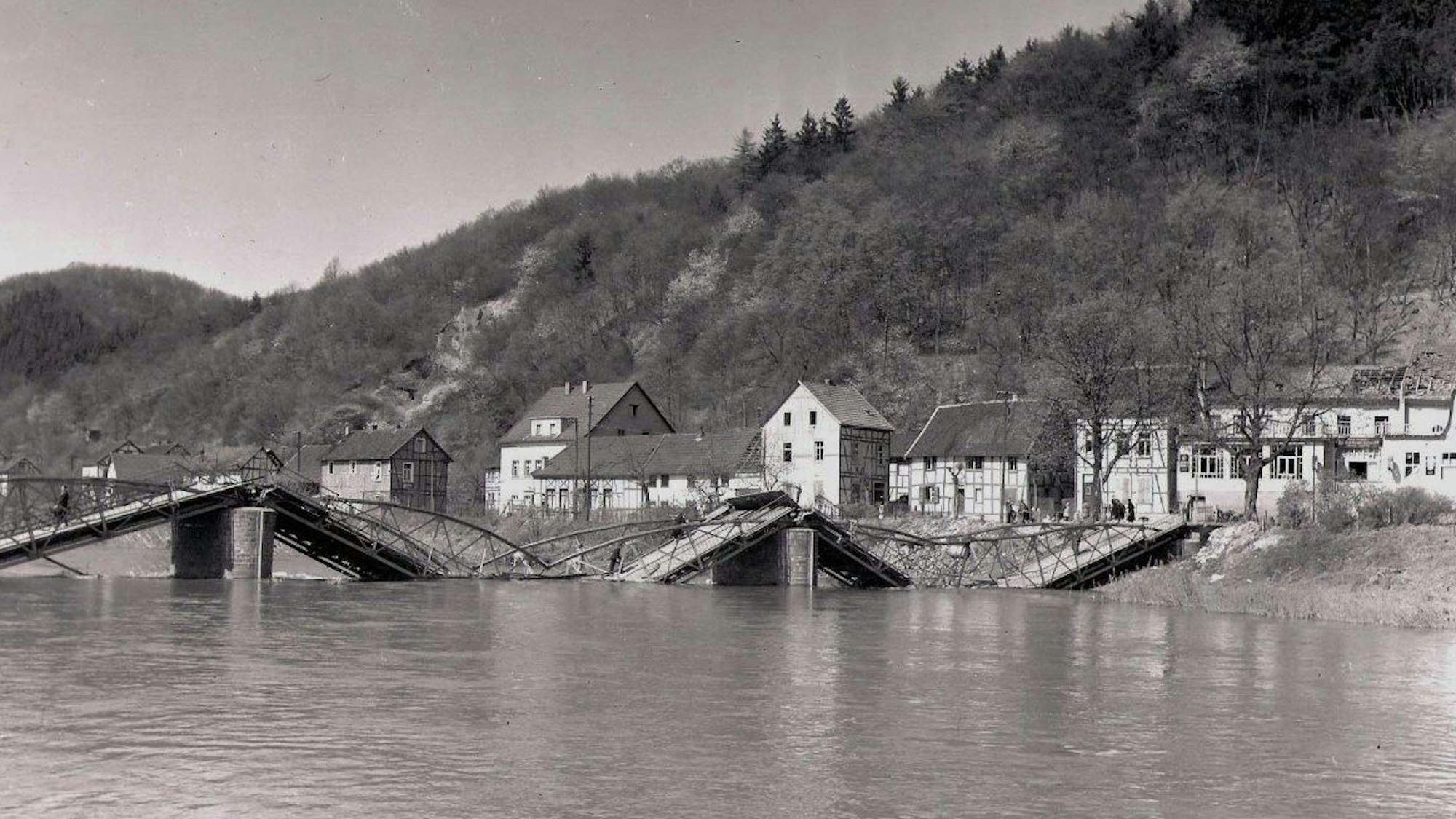 Schwarz-Weiß-Foto einer zerstörten Brücke; die Fahrbahnteile liegen im Fluss.