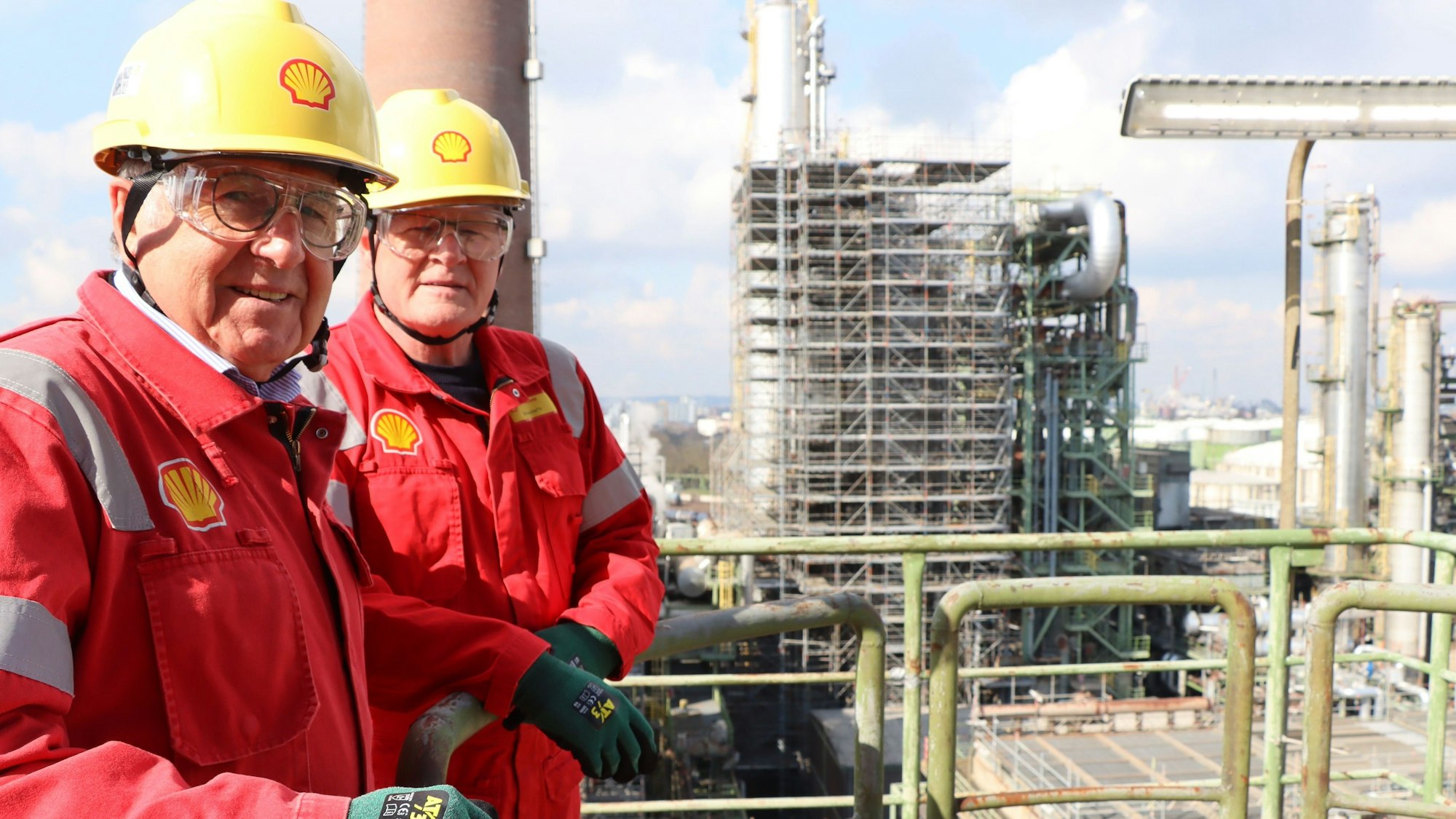 Zwei Männer in roten Anzügen und gelben Schutzhelmen stehen vor einer Produktionsanlage.