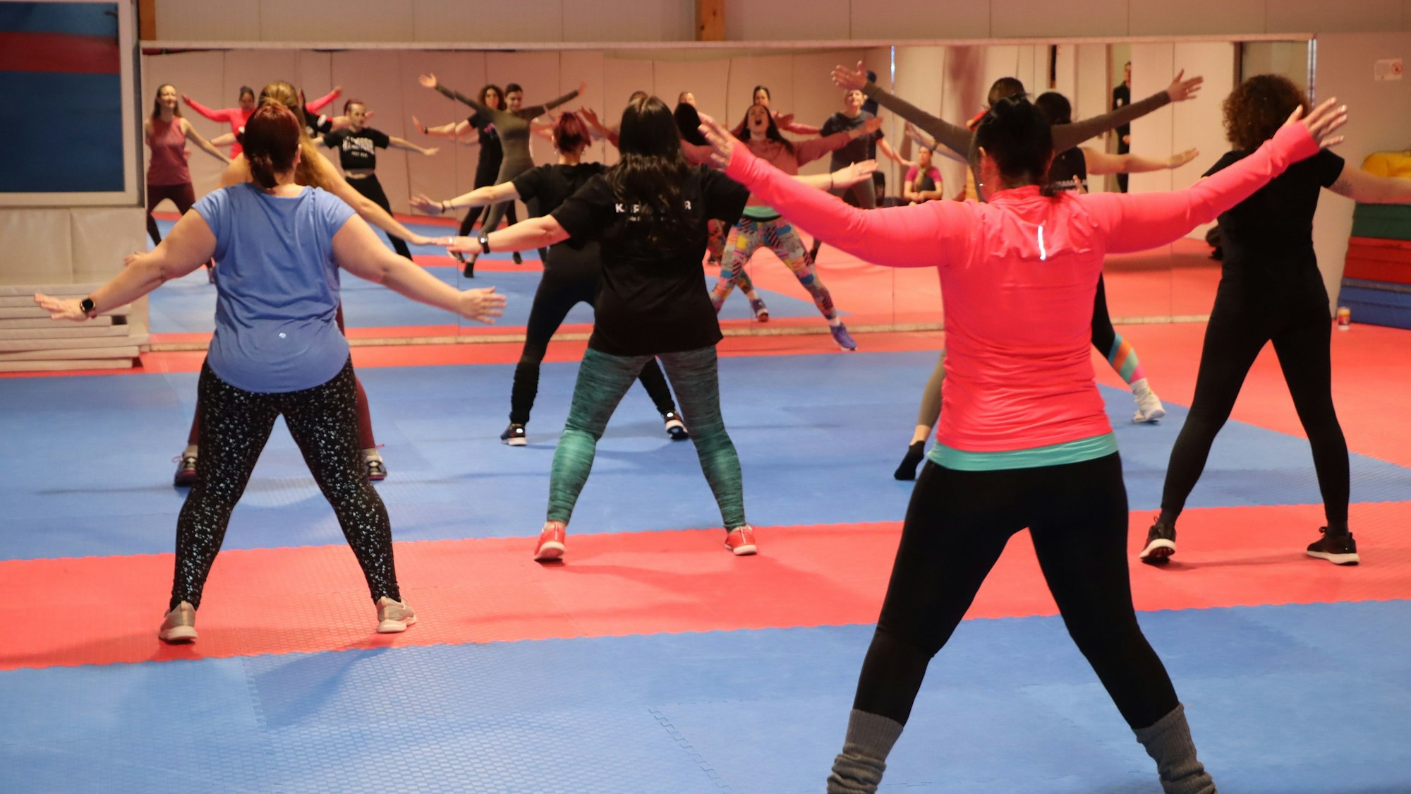 Zu sehen sind mehrere Frauen, die in Sportkleidung in einem Gymnastikraum vor einem Spiegel tanzen.