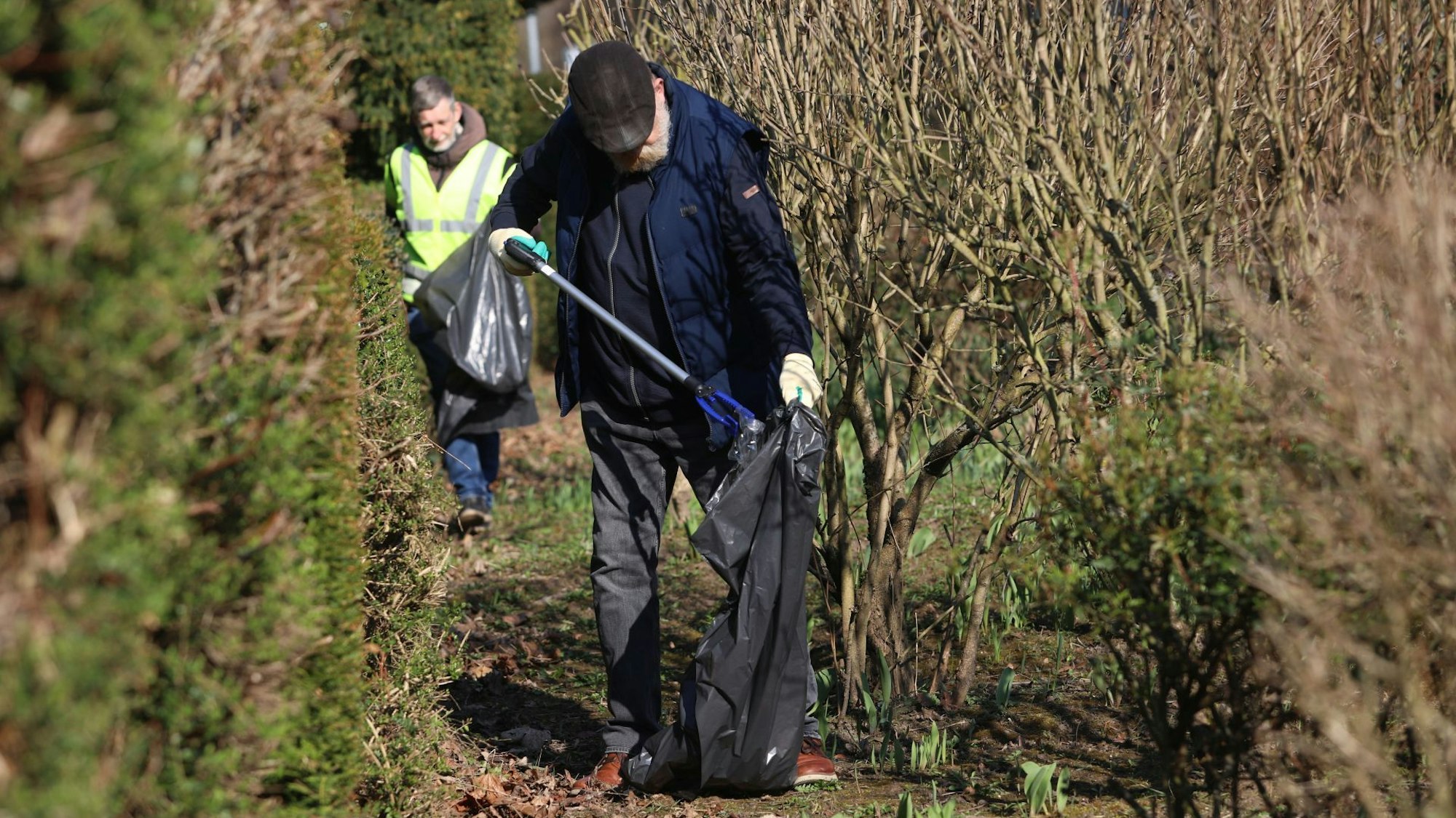 Zwei Männer sammeln Müll auf einem Grünstreifen, einer von ihnen legt mit seiner Zange gerade etwas in einen Müllbeutel.