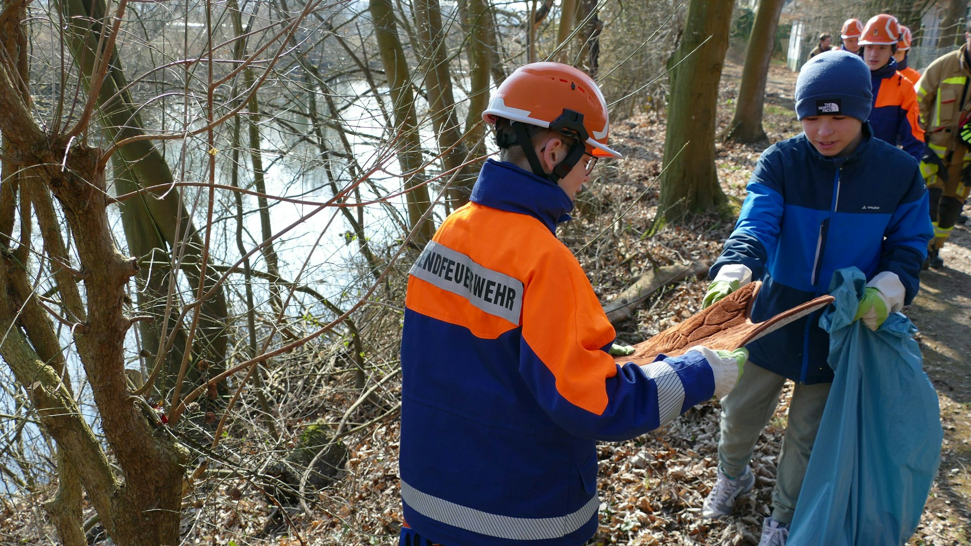 Zwei Jugendliche im Outfit der Siegburger Jugendfeuerwehr stehen an einer Böschung und halten eine Autofußmatte und einen blauen Müllsack in den Händen.