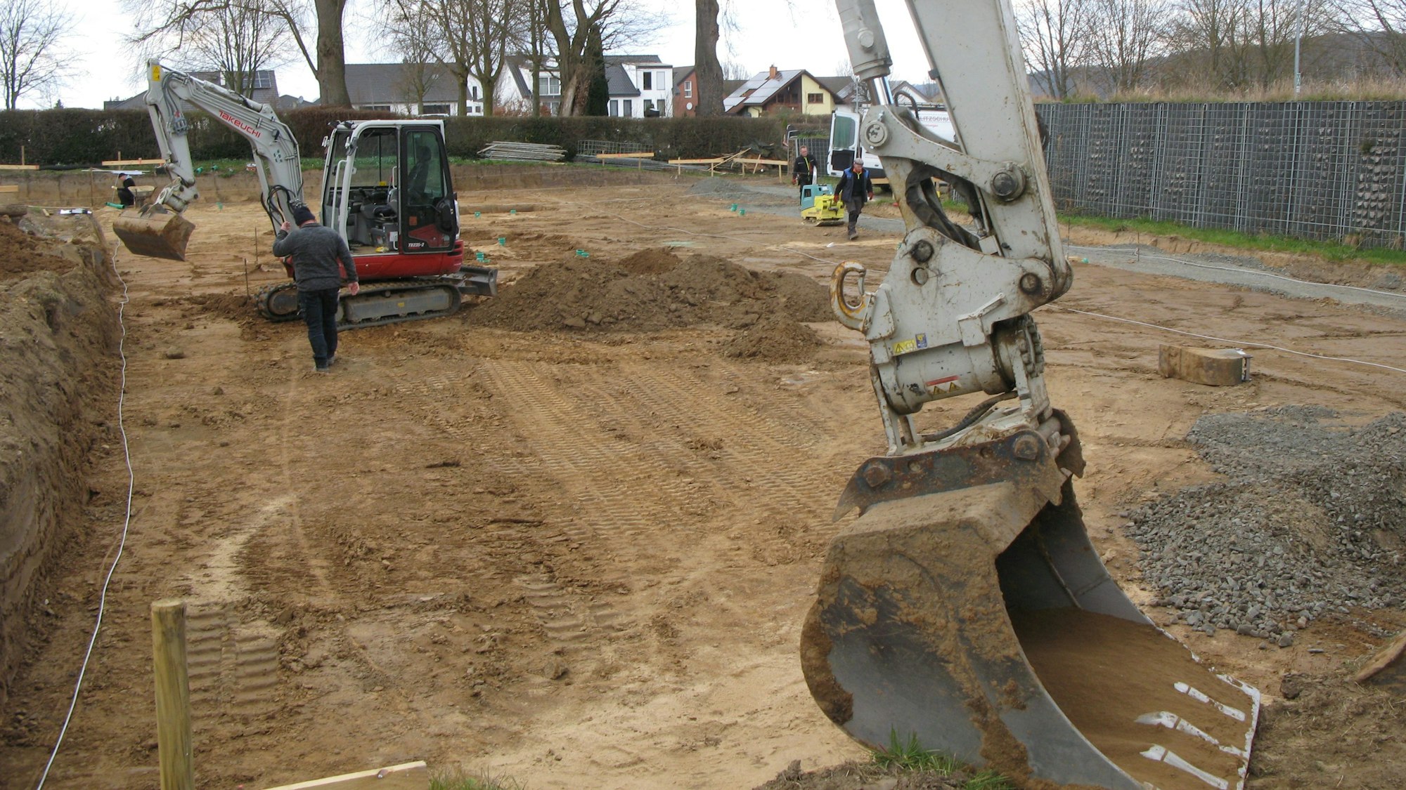Eine große Baggerschaufel im Vordergrund und ein kleiner Bagger in einer Baugrube.