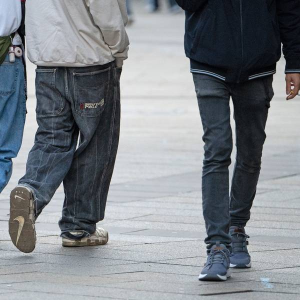 Drei Jugendliche in Jeans gehen durch die Innenstadt. Einer hält eine Zigarette in der Hand.