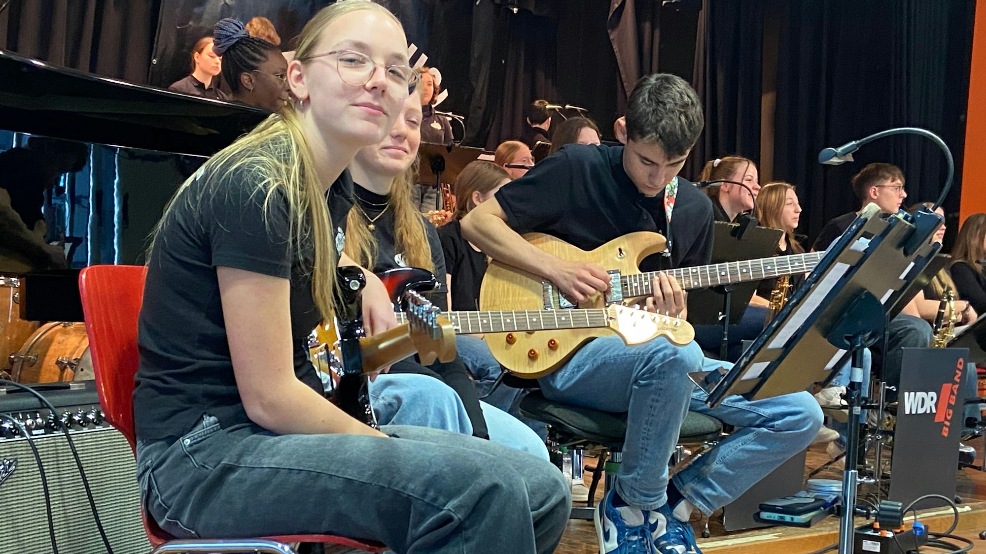 Ein Mädchen mit langem blonden Zopf und weitere Schülerinnen und Schüler sitzen auf einer Bühne.