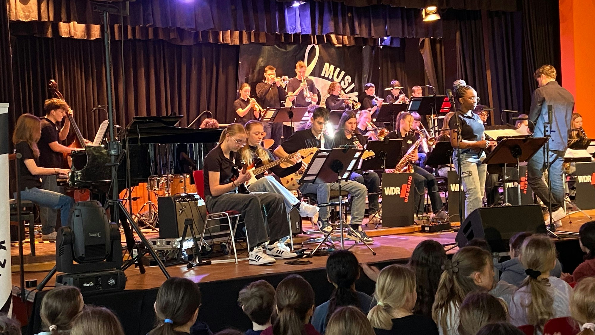 Auf einer Bühne ist ein Orchester zu sehen, im Publikum sitzen viele Schüler des Porzer Stadtgymnasiums.