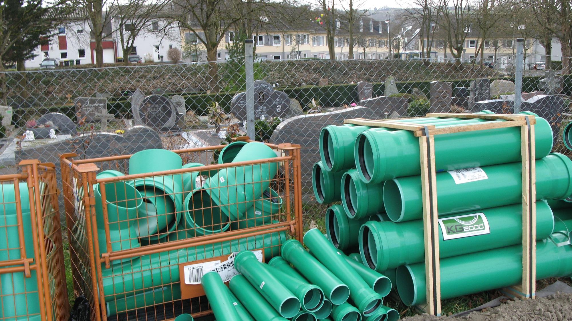 Grüne Baurohre lagern vor einem Zaun, hinter dem Grabsteine eines Friedhofs zu sehen sind.