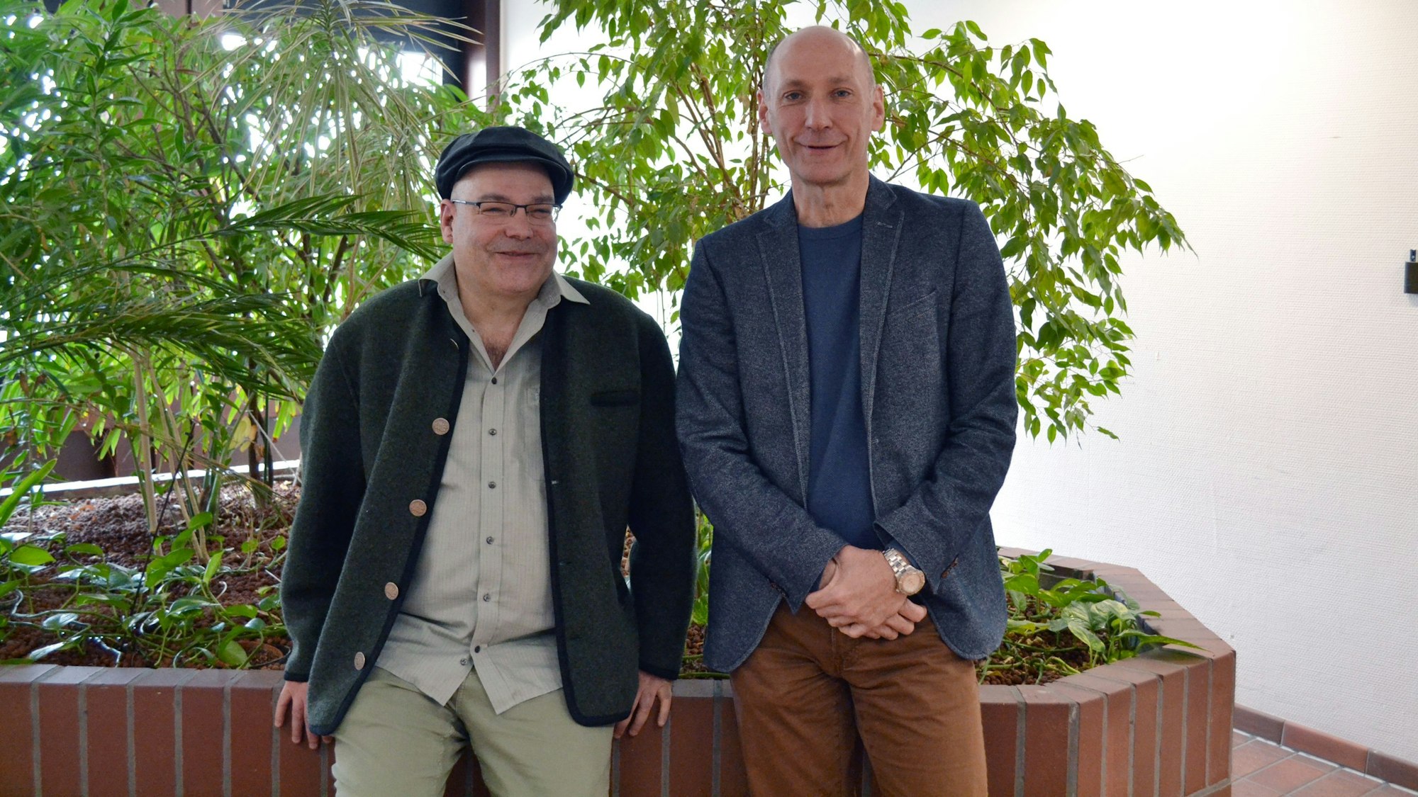 Zwei Männer stehen vor grünen Pflanzen, die von einer Backsteinmauer umgeben sind.