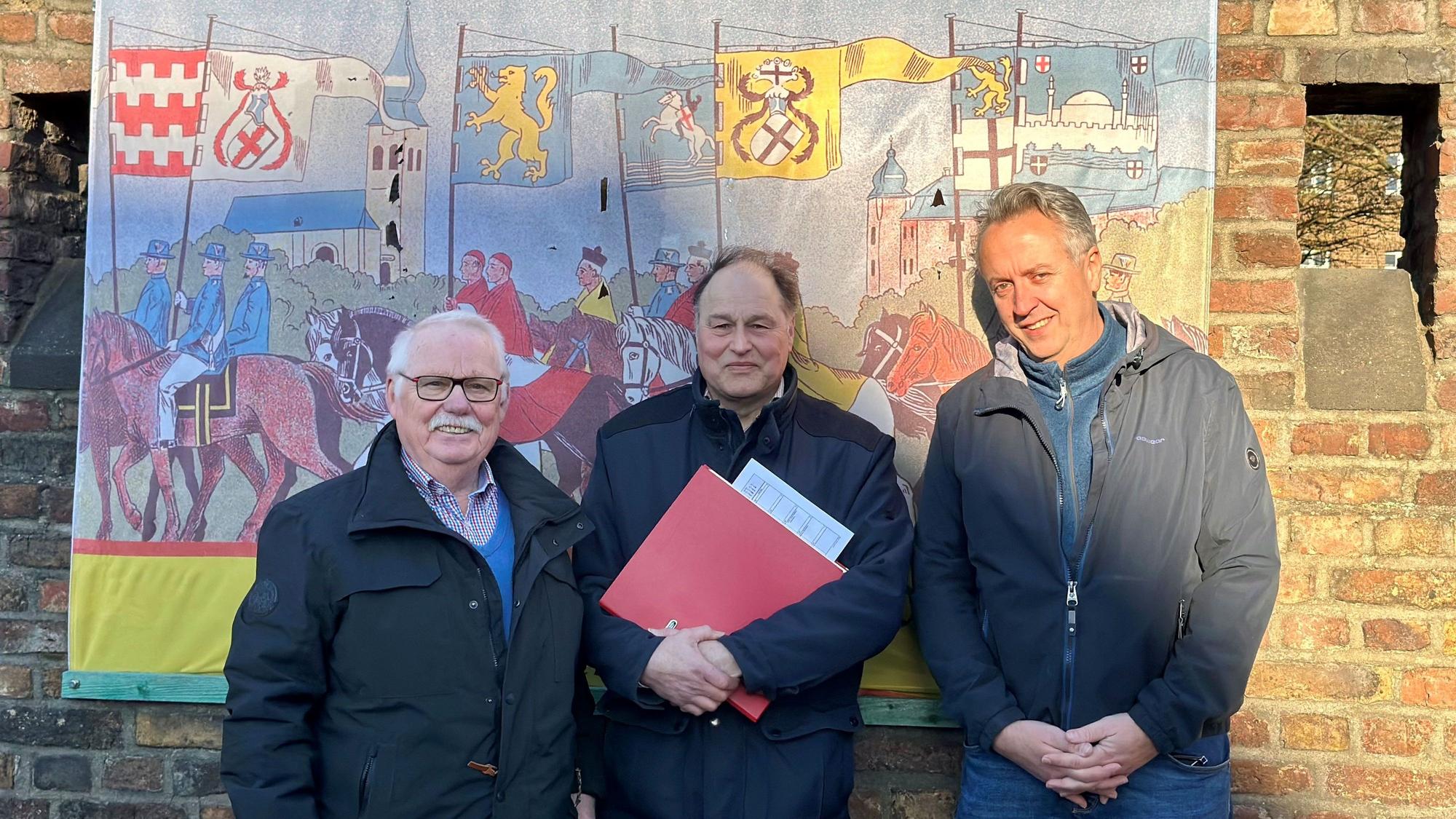 Drei Männer stehen vor einem Banner, das an einer Backsteinwand hängt.