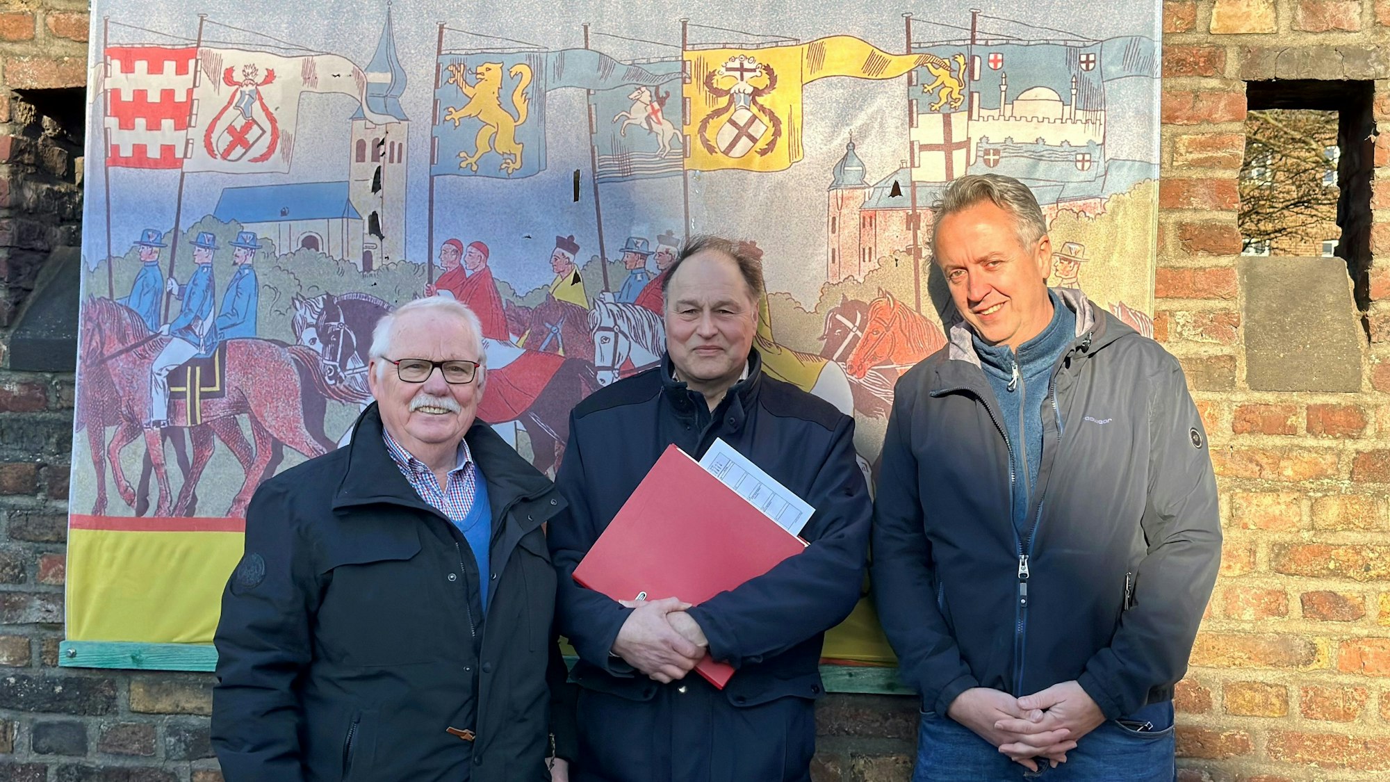 Drei Männer stehen vor einem Banner, das an einer Backsteinwand hängt.